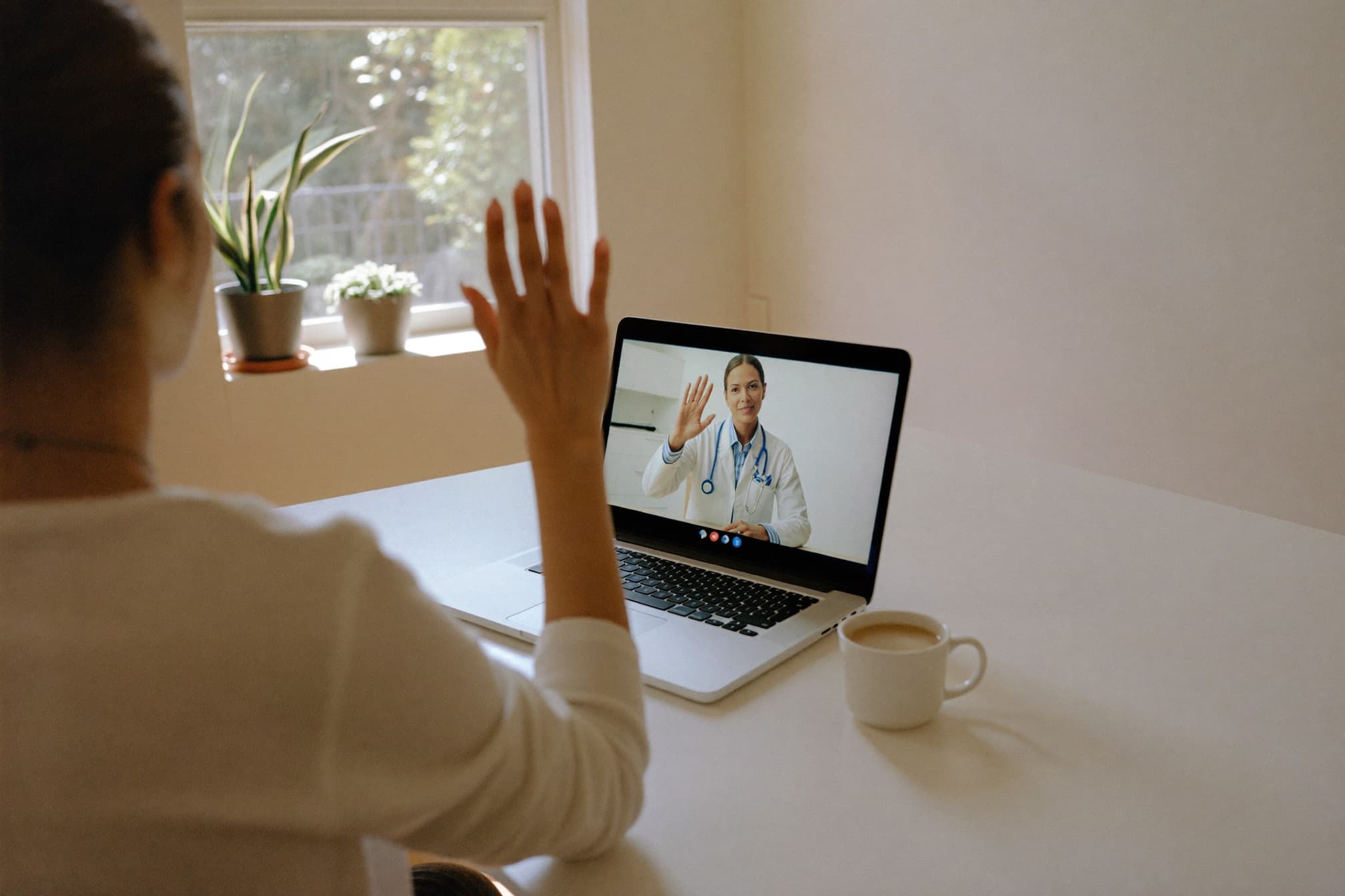 A woman at her kitchen table waves to her clinician during a video visit